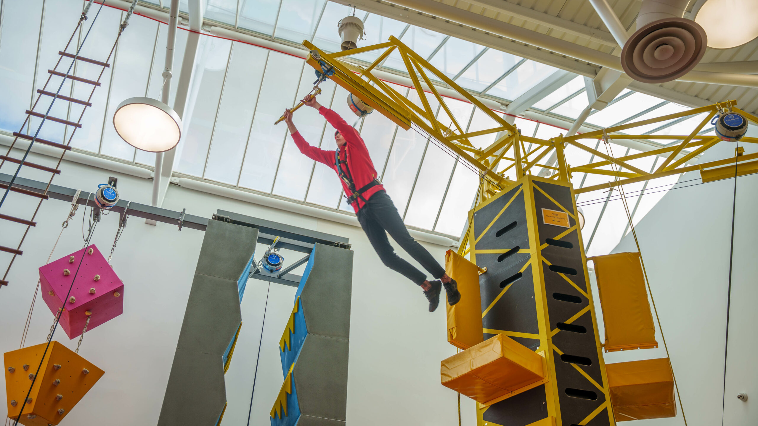 Person in red shirt climbing a high ropes course in an indoor adventure park with geometric obstacles.