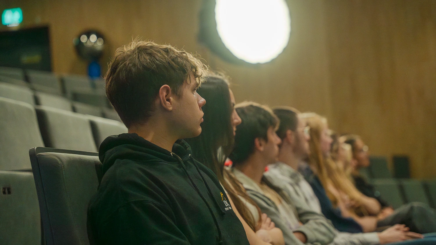 Audience attentively watching a presentation in a theater, with focus on a young man in a hoodie.