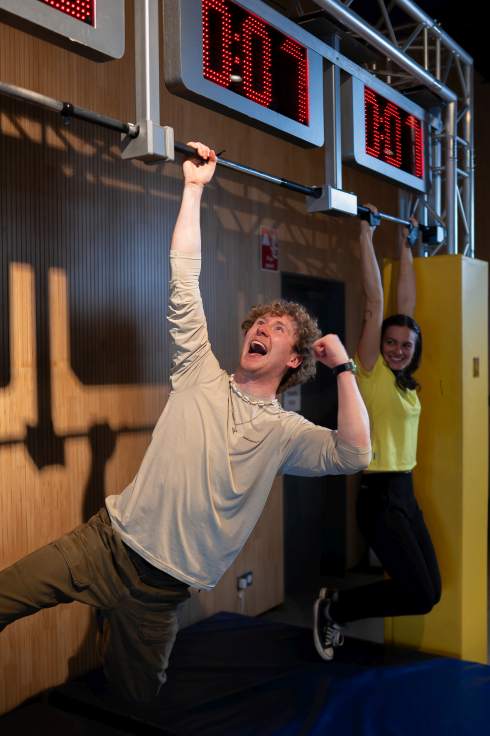 Two people competing on monkey bars indoors, one cheering with effort, race timer in the background.